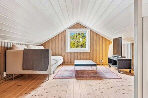 Ein gemütliches Dachzimmer mit schrägen Wänden, einer Couch, einem kleinen Tisch, einem Fernseher und einem Fenster mit Blick auf Bäume.