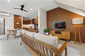 A modern living room with a wooden sofa, a dining area with a table and chairs, a kitchen with wooden cabinets, and a mounted television.