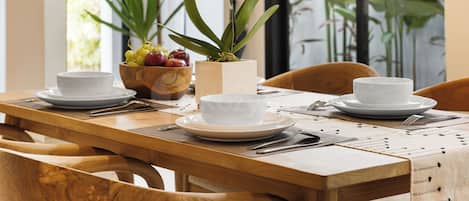 Une table de salle Ă manger en bois, avec des assiettes blanches, un saladier de fruits et une plante en pot.