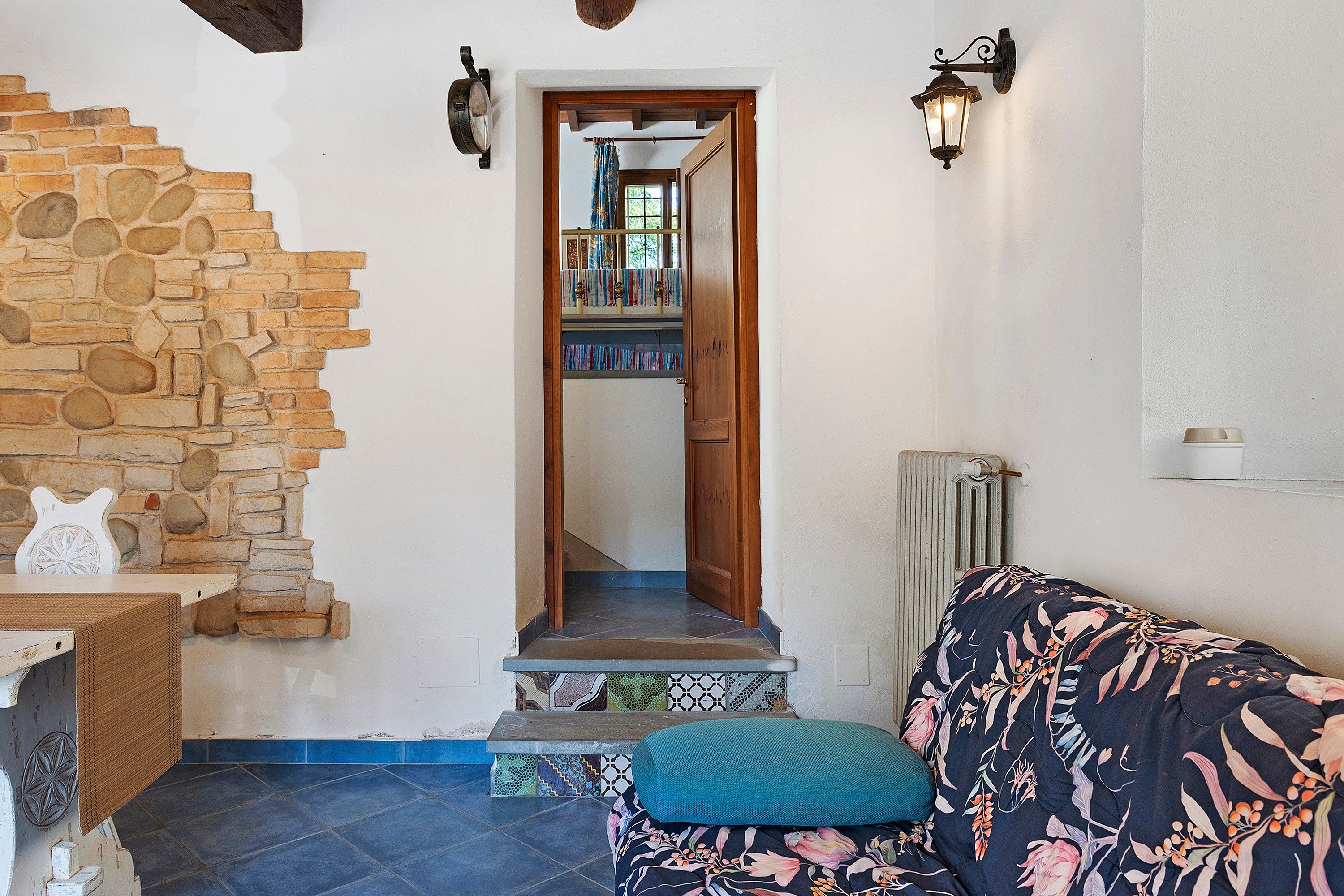 Une pièce avec un mur en pierre, une porte en bois donnant sur un escalier, un canapé à motifs et un radiateur.