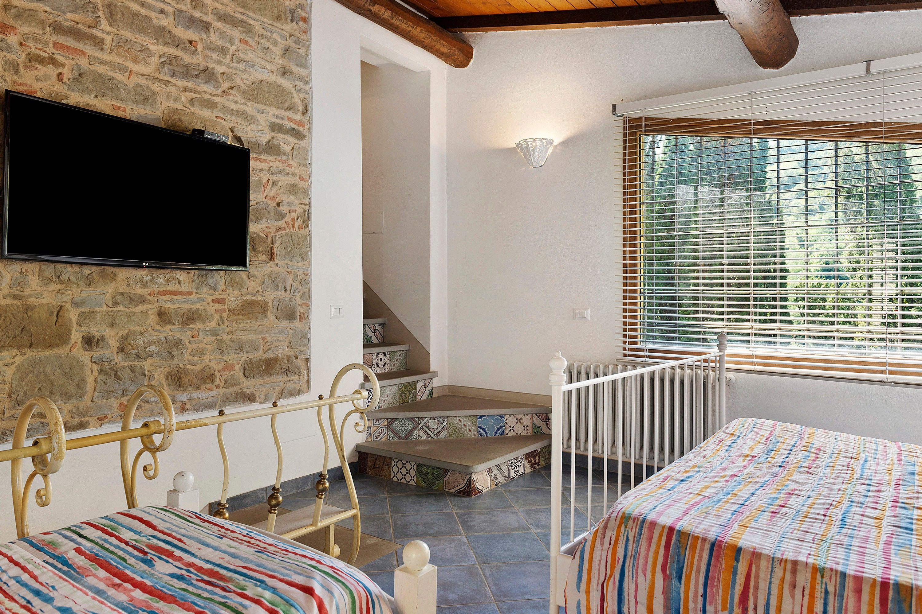 Habitació amb paret de pedra, un televisor de pantalla plana fixat a ella, escala amb rajoles estampades, finestra amb persianes i llit amb llençols a ratlles.