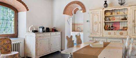 A rustic kitchen with exposed brick arches, a white cabinet with glassware, and a dining table with chairs.