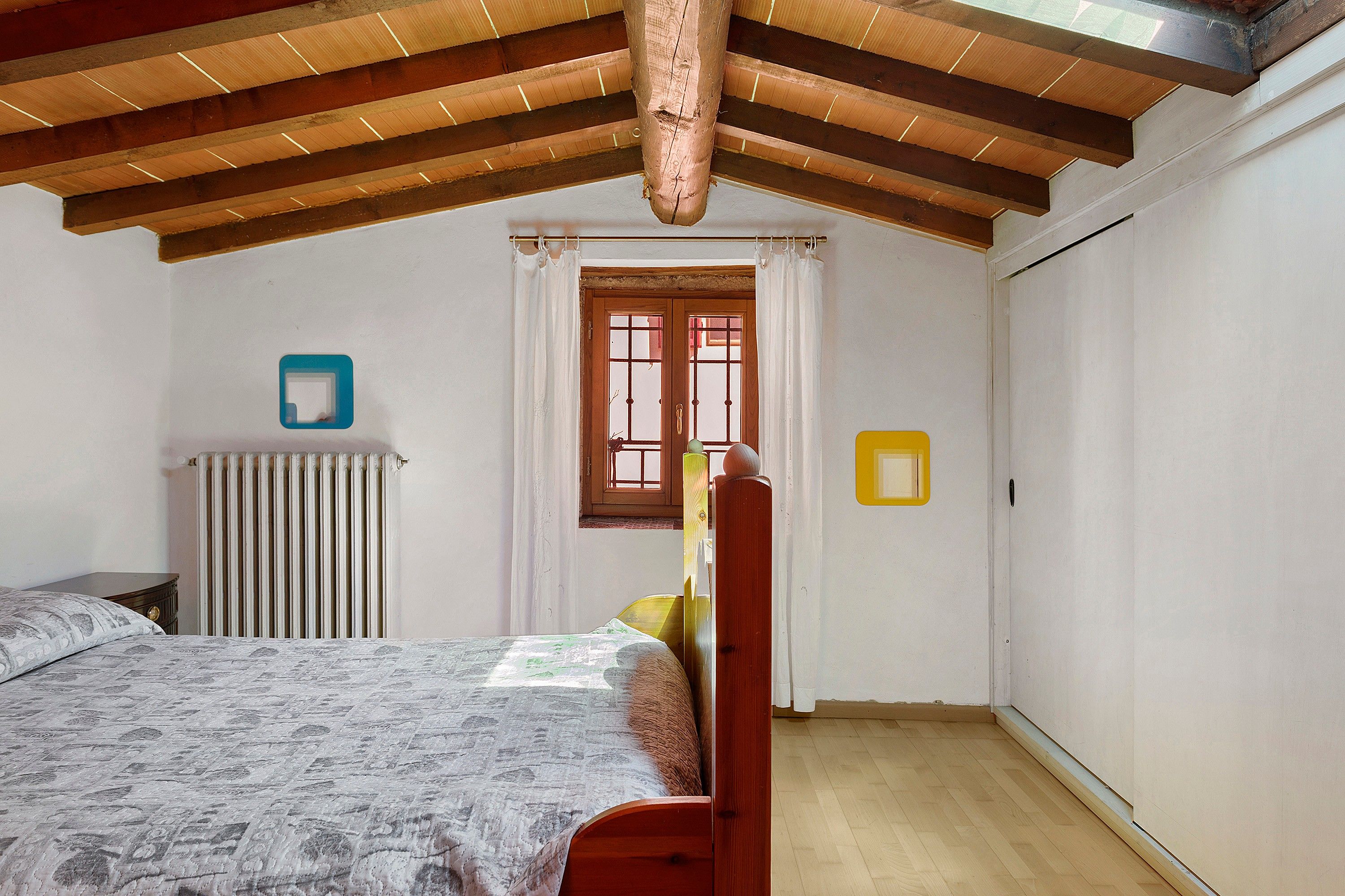 A bedroom with a wooden ceiling, a radiator, a bed with a patterned bedspread, a window with curtains, and a door.