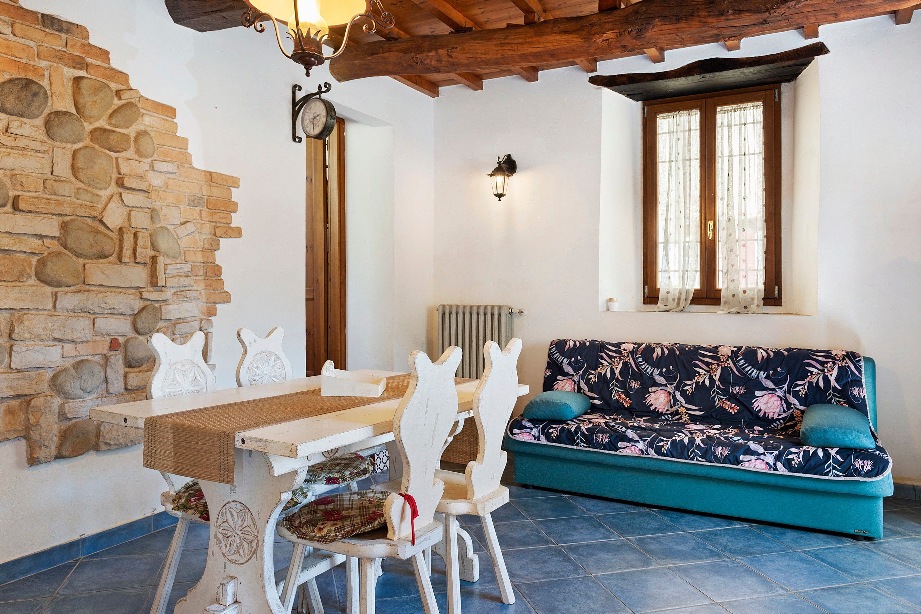 A cozy room with a stone wall, a wooden ceiling, a dining table with chairs, a sofa with floral upholstery, and a window with curtains.