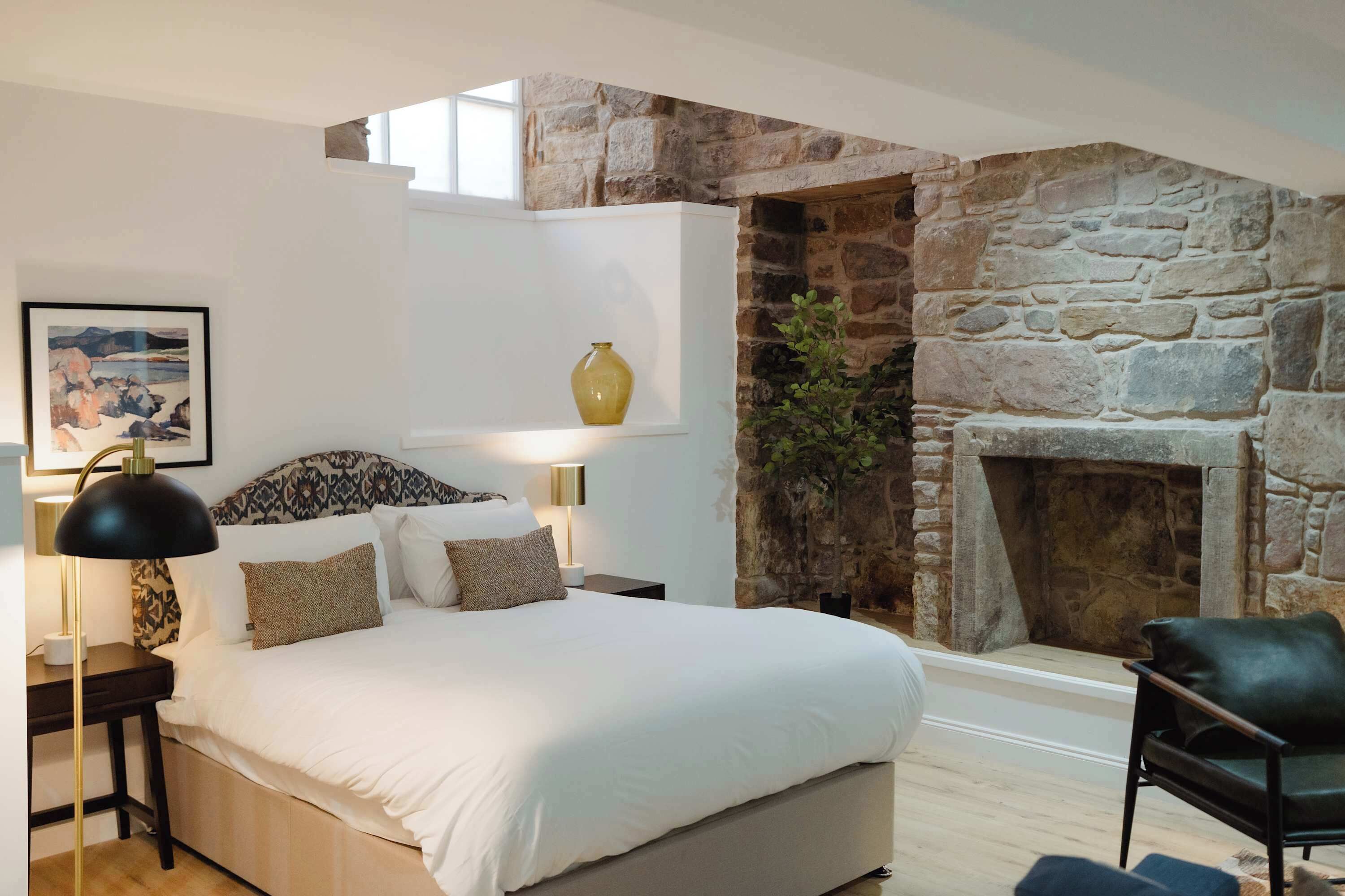 A bedroom with a stone fireplace, a bed with white bedding, a nightstand with a lamp, and a framed picture on the wall.