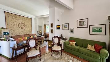 A living room with a green sofa, wooden chairs, a dining table, and a large tapestry.