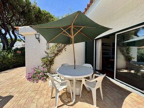 Een terras buiten met een ronde tafel, twee stoelen, een groene parasol en een witte muur met een raam.