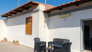 A building with a tiled roof, a window with a shutter, and a door leading to an interior space. There are two wicker chairs and a small table with a potted plant. - CASA SAURO 10 (Avola)