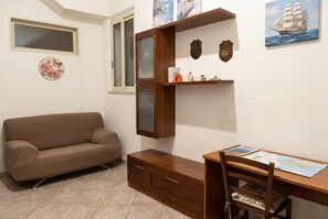 Ein Zimmer mit einem braunen Sofa, einem Holztisch mit Stuhl, einem Fenster und Wanddekorationen, darunter ein Gemälde eines Schiffes und ein Wappen.