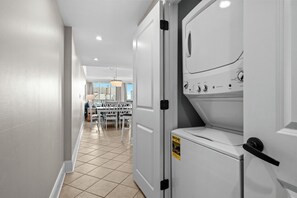 A laundry room with a washing machine and dryer, a tiled floor, and a view into a dining area with chairs and a table. - 10% off 3-7 nights thru Dec. Ocean Drive. Oceanfront with OCEAN VIEW (from side). (North Myrtle Beach)