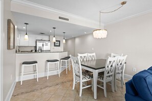 A dining area with a table and chairs, a kitchen in the background, and a hanging light fixture. - 10% off 3-7 nights thru Dec. Ocean Drive. Oceanfront with OCEAN VIEW (from side). (North Myrtle Beach)