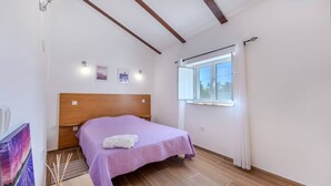 Ein Schlafzimmer mit einem hölzernen Kopfteil, einem Bett mit violetter Bettdecke, einem Fenster mit Vorhängen und einem kleinen Nachttisch.