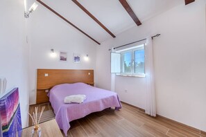 Ein Schlafzimmer mit einem hölzernen Kopfteil, einem Bett mit violetter Bettdecke, einem Fenster mit Vorhängen und einem kleinen Nachttisch.
