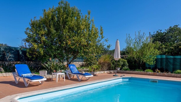 Ein Swimmingpool mit zwei Liegestühlen und einem Tisch, umgeben von Grünflächen und wolkenlosem Himmel.