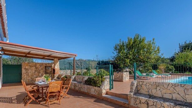 Um pátio com mesa e cadeiras, um muro de pedra, uma piscina e uma cerca verde.