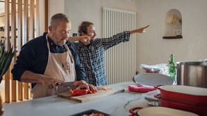 Dos personas en una cocina con tabla de cortar, verduras y utensilios de cocina.