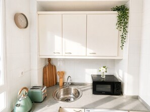 Una cocina con gabinetes blancos, un lavamanos de acero inoxidable, microondas, tostadora, hervidor de agua, utensilios de madera y una planta en maceta.