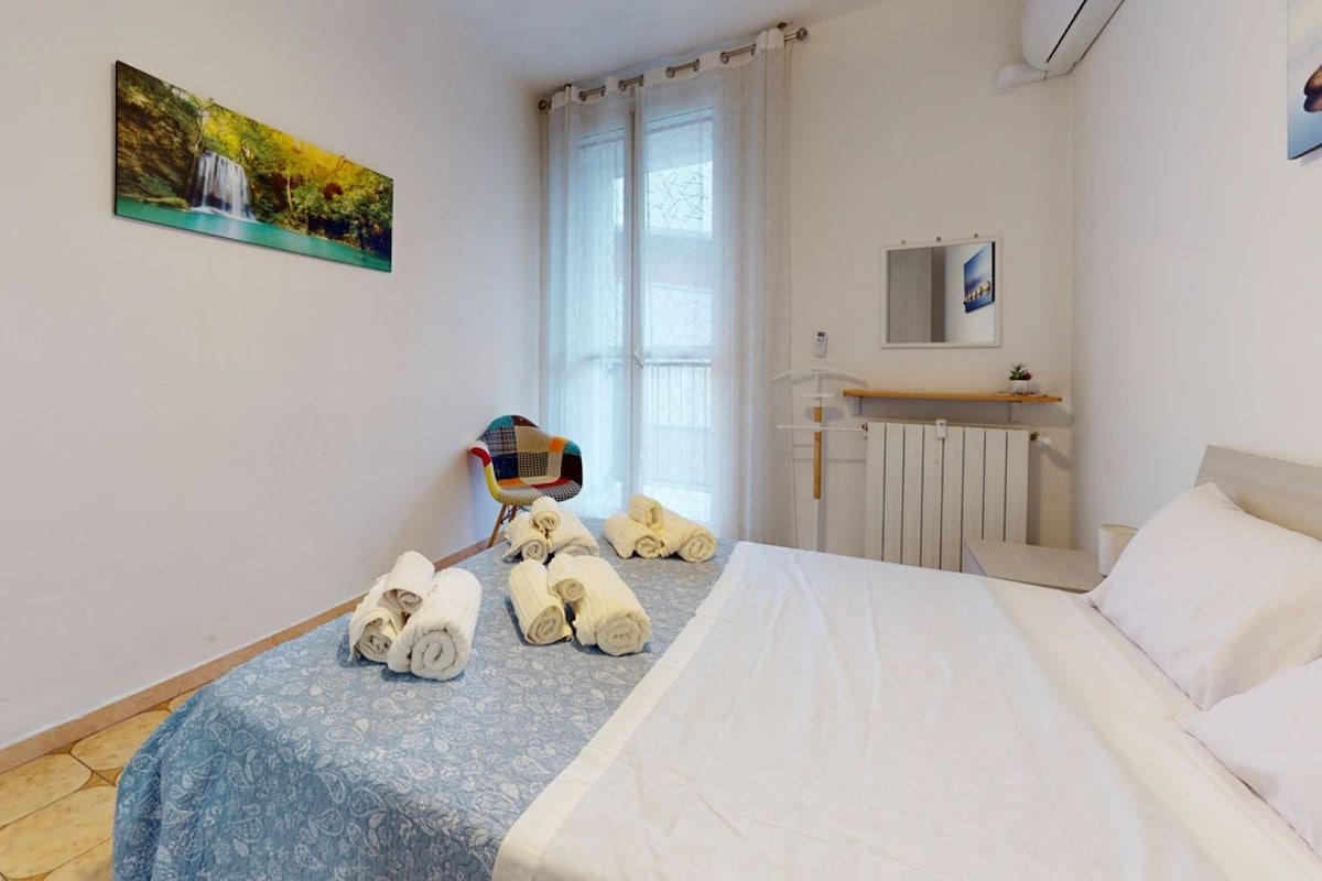 A hotel room with a bed, a chair, a radiator, and a window with curtains.