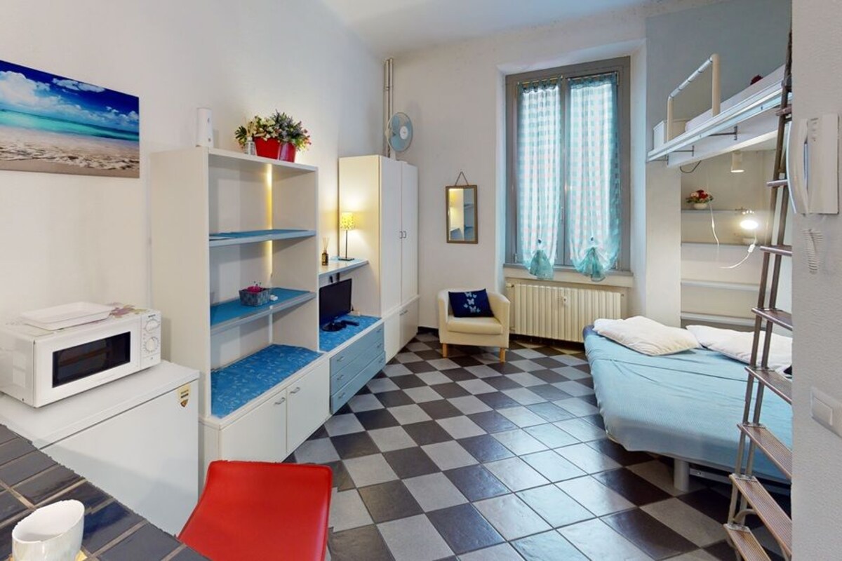A compact room with a checkered floor, a bed, a desk with a computer, a microwave, and a window with curtains.