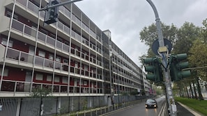 A multi-story residential building with balconies, a pedestrian walkway, and a street with traffic lights.