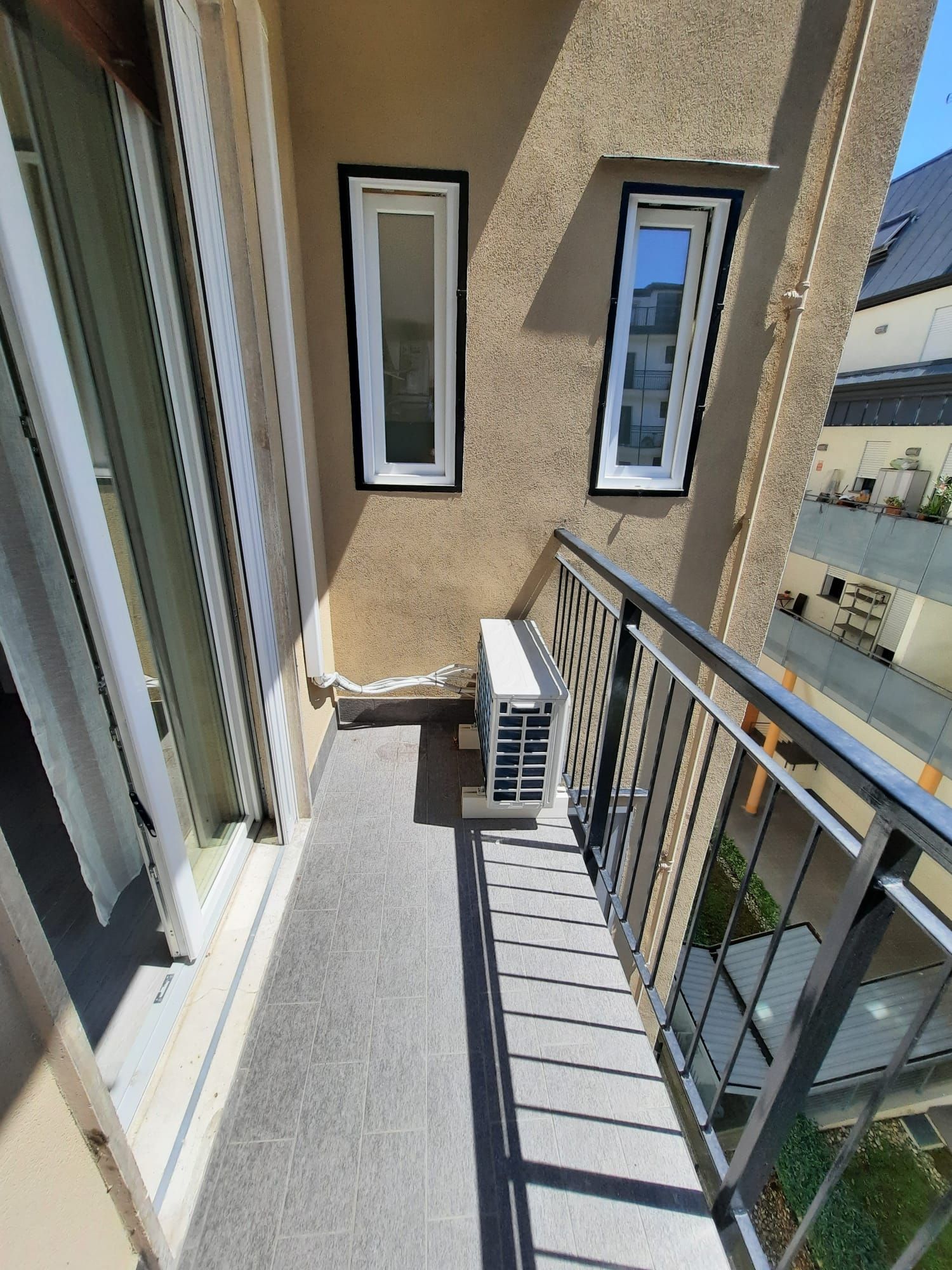 Un balcon avec une rambarde en métal, un petit climatiseur et deux fenêtres à cadre blanc.
