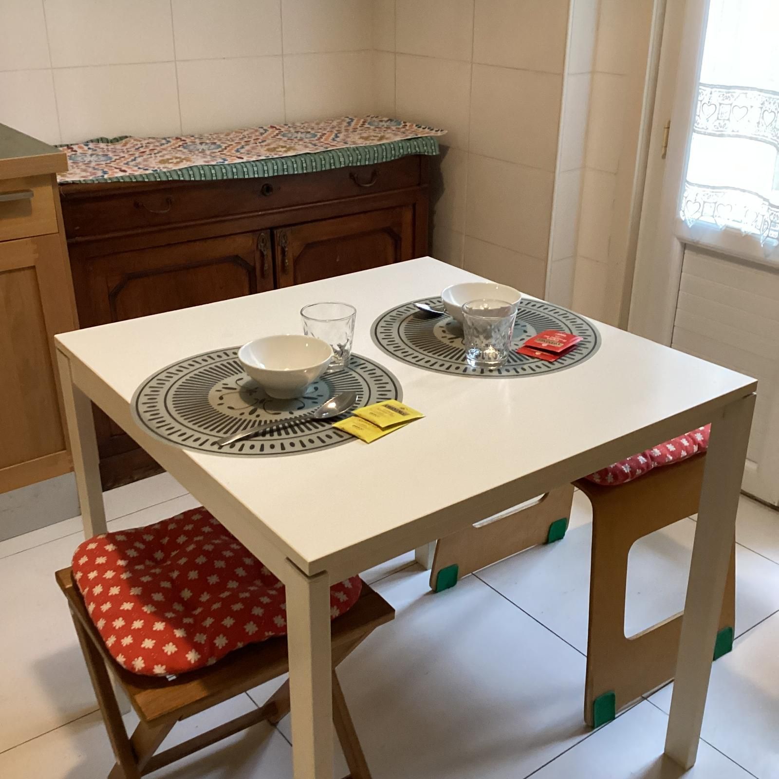 A small dining table with two plates, two glasses, and a spoon. There is a chair with a red cushion.