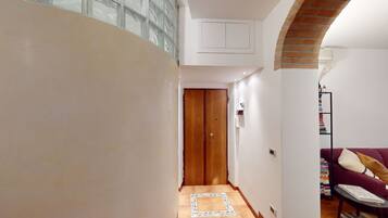 Un pasillo estrecho con puerta de madera, suelo de baldosas, estantería y un sofá rojo.