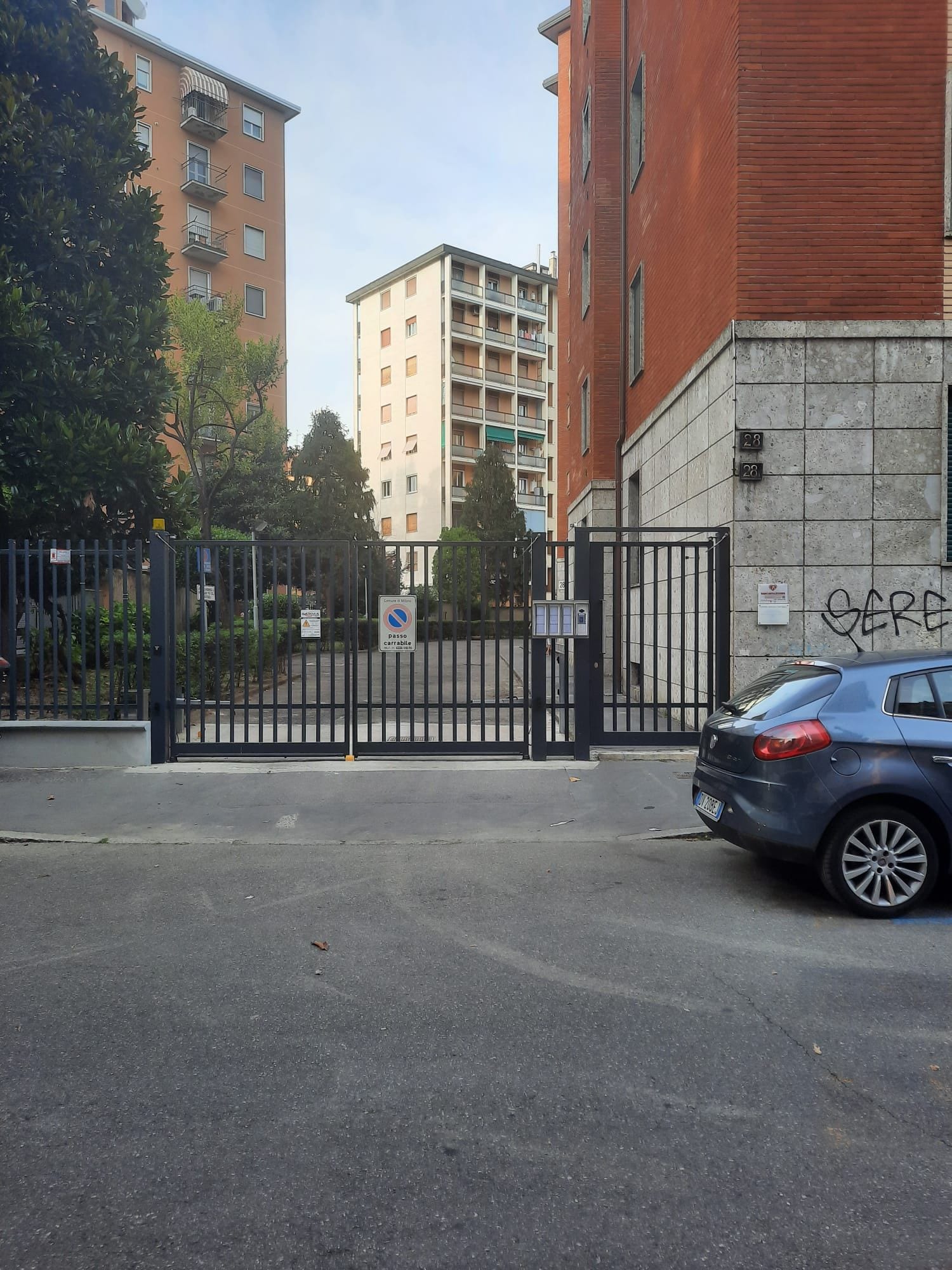 Une entrée fermée par un portail donnant accès à un quartier résidentiel, avec une voiture bleue garée à proximité.