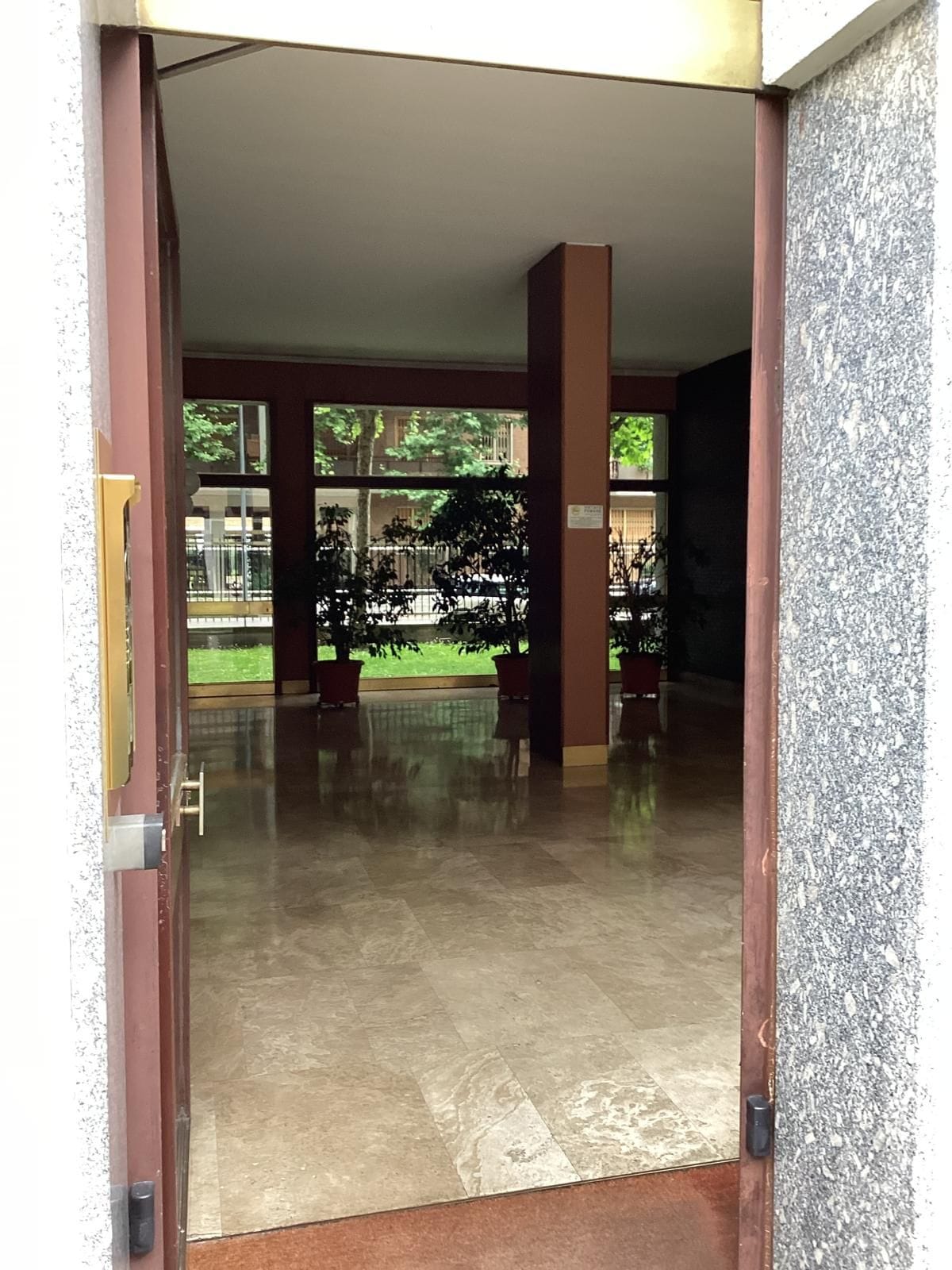 An open door leading to a spacious lobby with a tiled floor and a view of a courtyard with trees and a fence.