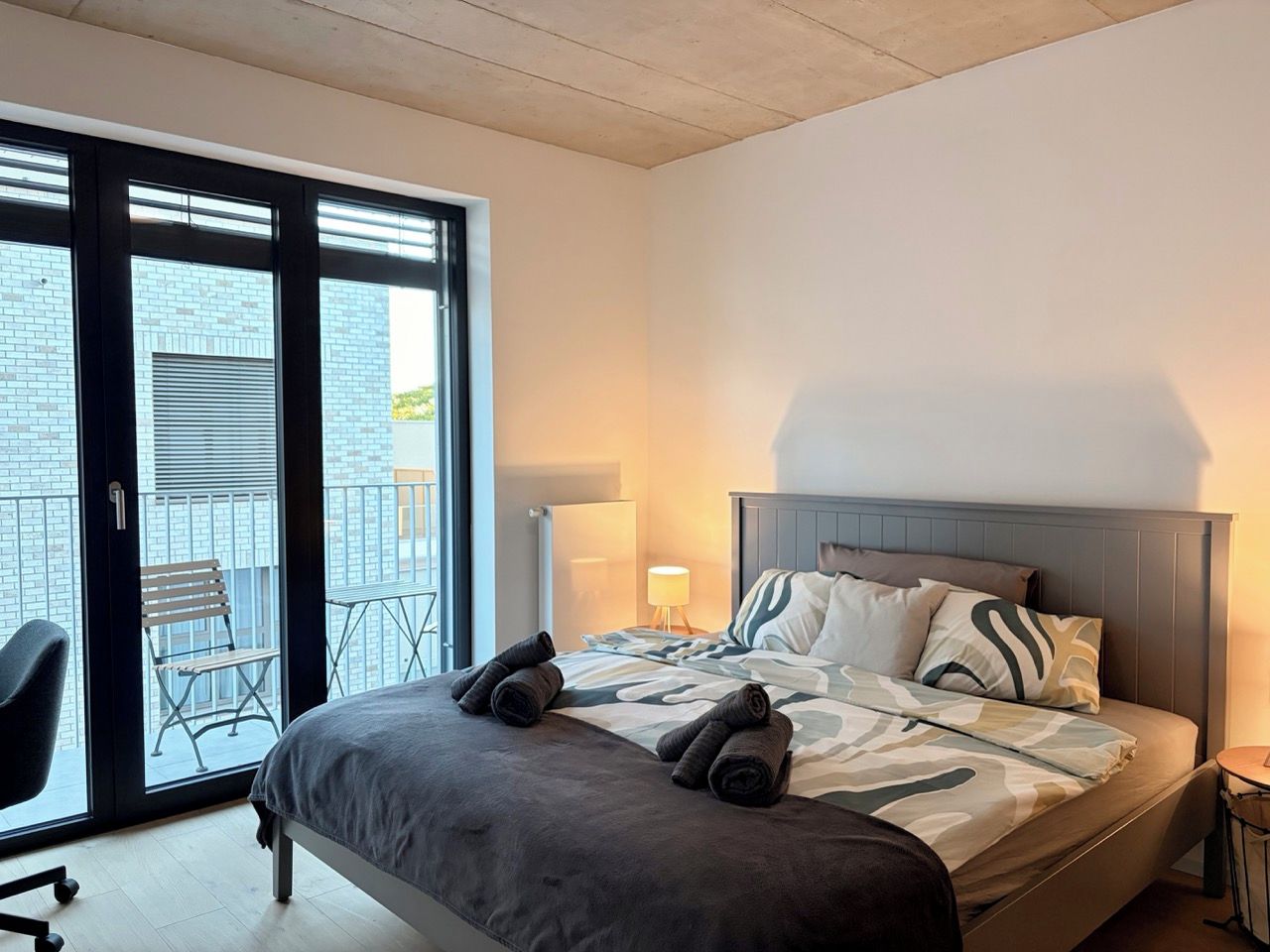 A modern hotel room with a bed, a chair, a lamp, and a balcony with a chair.