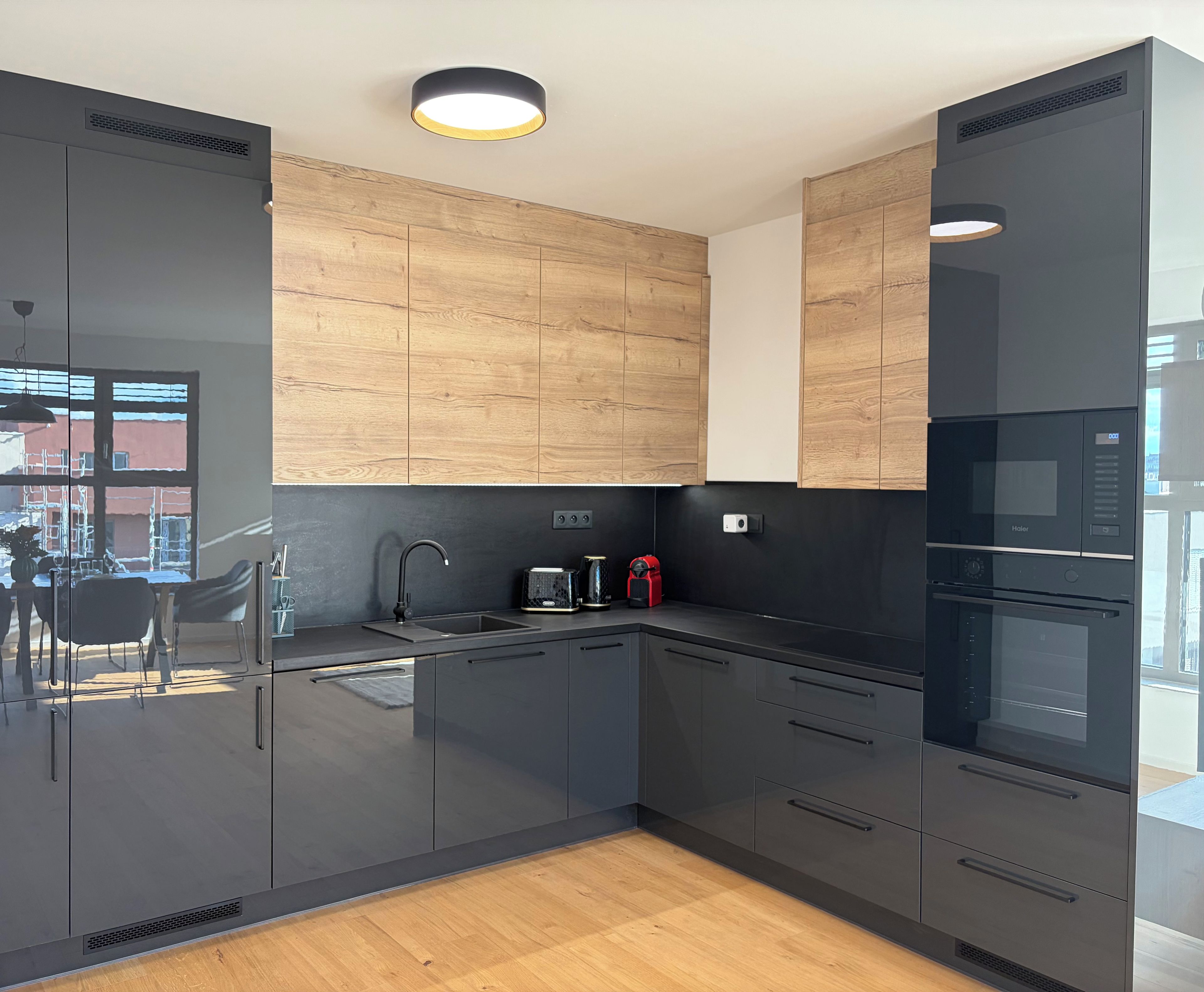 A modern kitchen with dark cabinetry, a wooden accent wall, a built-in oven, and a window with a view of the outside.