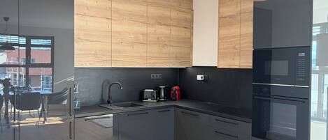 A modern kitchen with dark cabinetry, a wooden accent wall, a built-in oven, and a window with a view of the outside.