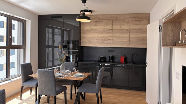 A modern kitchen with dark cabinetry, a wooden countertop, and a dining area with grey chairs and a wooden table.