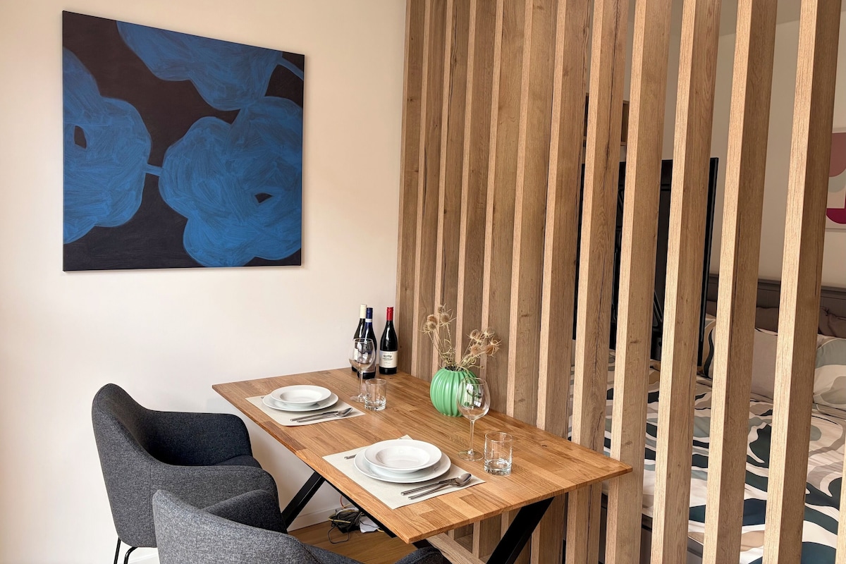 A dining area with a wooden table set for two, a modern chair, a wall-mounted abstract painting, and a wooden partition.