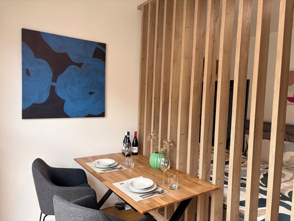 A dining area with a wooden table set for two, a modern chair, a wall-mounted abstract painting, and a wooden partition.