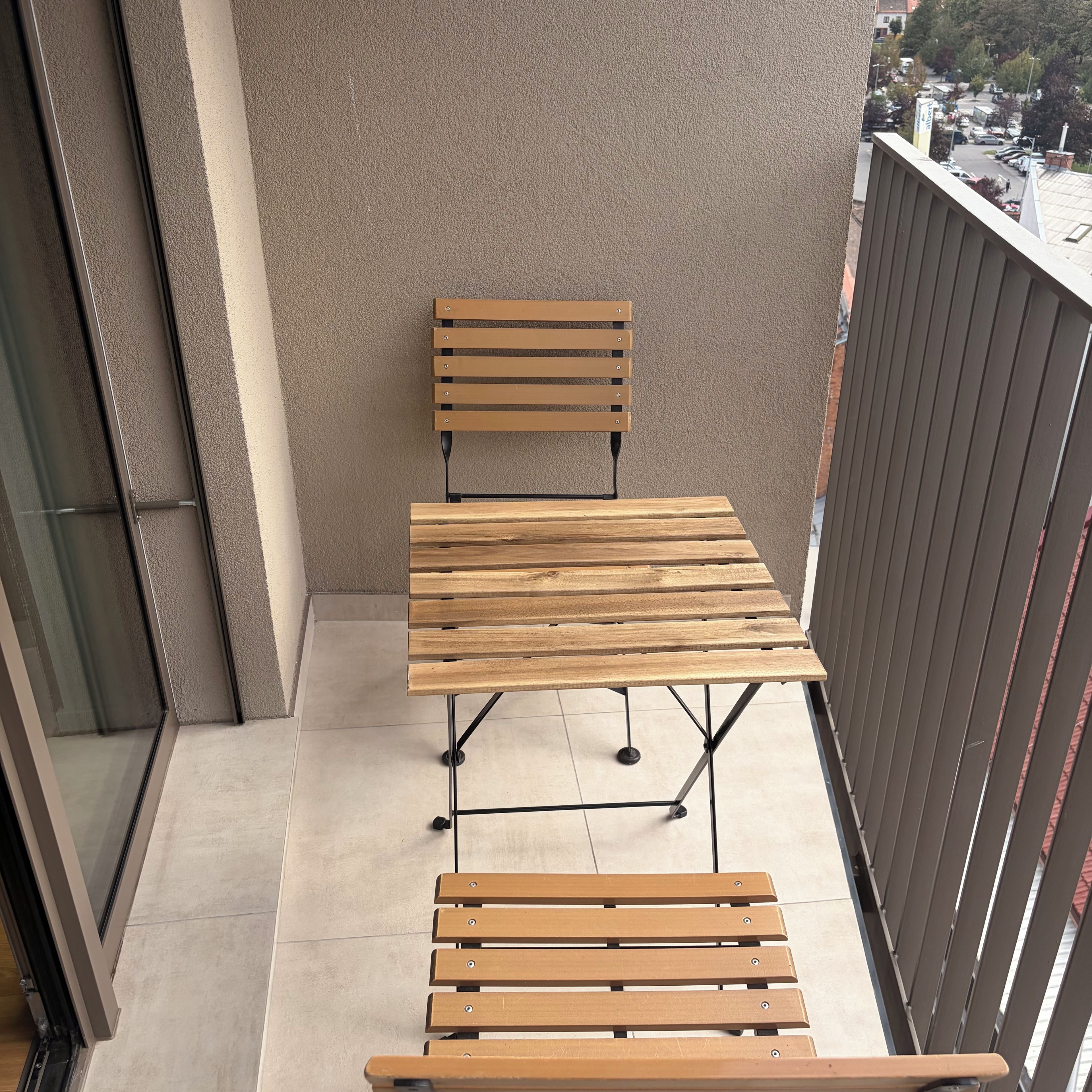 A balcony with a wooden table and bench, a view of the street, and a building in the background.