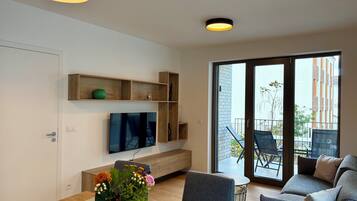 A modern living room with a flat-screen TV, wooden shelves, a dining table with a vase of flowers, and a sliding glass door leading to an outdoor area.