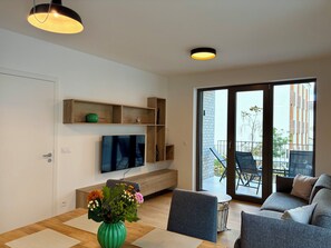 A modern living room with a flat-screen TV, wooden shelves, a dining table with a vase of flowers, and a sliding glass door leading to an outdoor area.