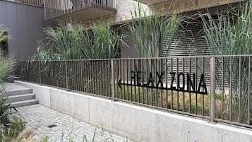 A gated entrance with a sign reading 'REXTONA' and a concrete pathway leading to a building with a metal railing and vegetation.