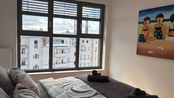 A room with a large window, a bed with a patterned bedspread, a painting on the wall, and a view of buildings outside.