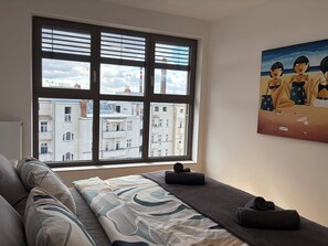 A room with a large window, a bed with a patterned bedspread, a painting on the wall, and a view of buildings outside.