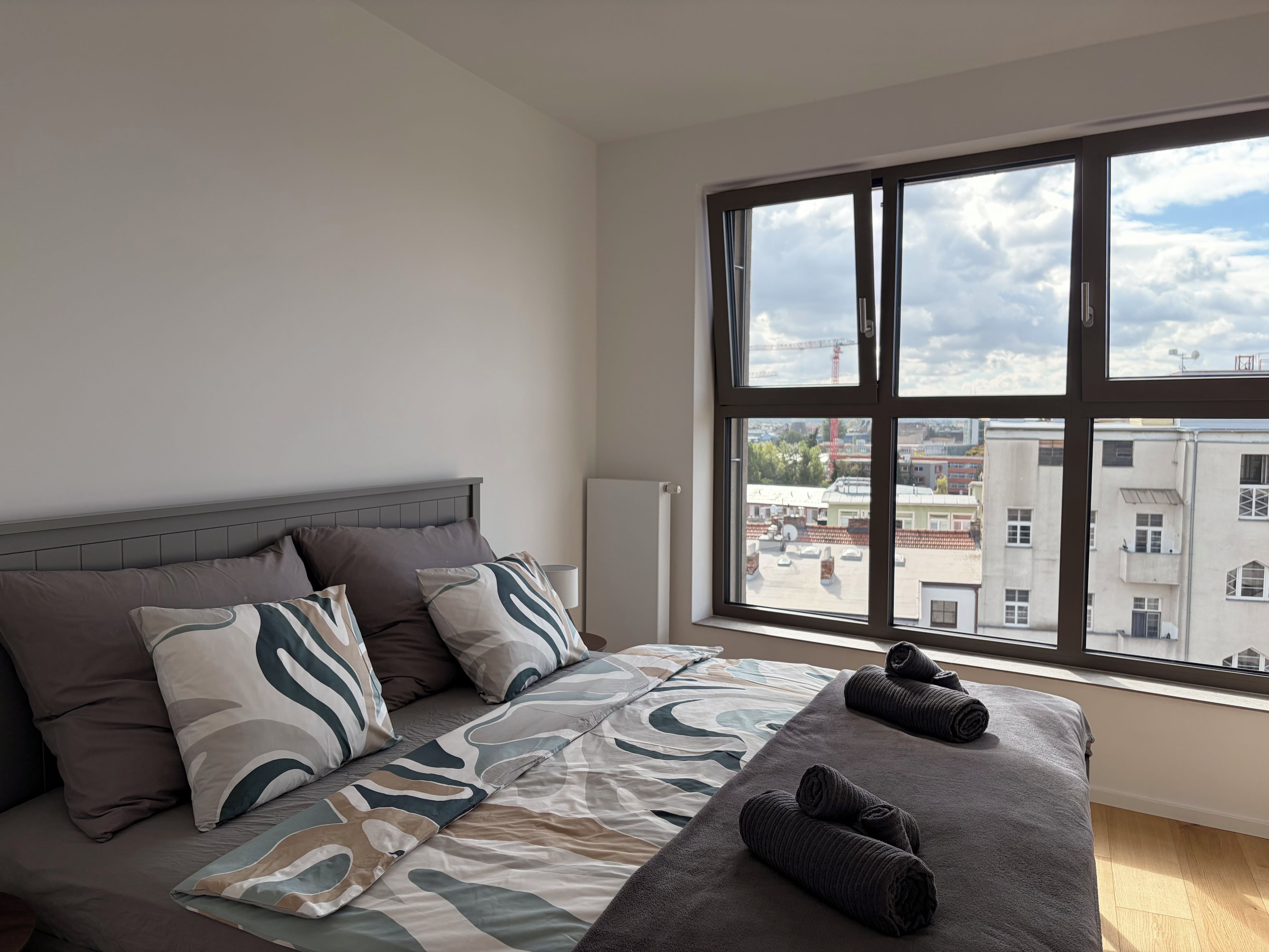 Een slaapkamer met een groot raam, een bed met beddengoed met een patroon, en uitzicht op gebouwen.