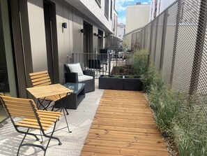 Un patio moderno al aire libre con cubierta de madera, una mesa con cubierta de vidrio, muebles de mimbre y una cerca de metal.