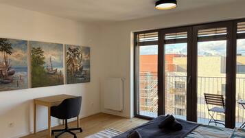 A bedroom with a bed, a desk, a chair, and a view of the city through large windows.