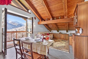 Uma sala de jantar com mesa posta para quatro, uma cozinha com armários de madeira e vista para montanhas cobertas de neve através de uma grande janela.