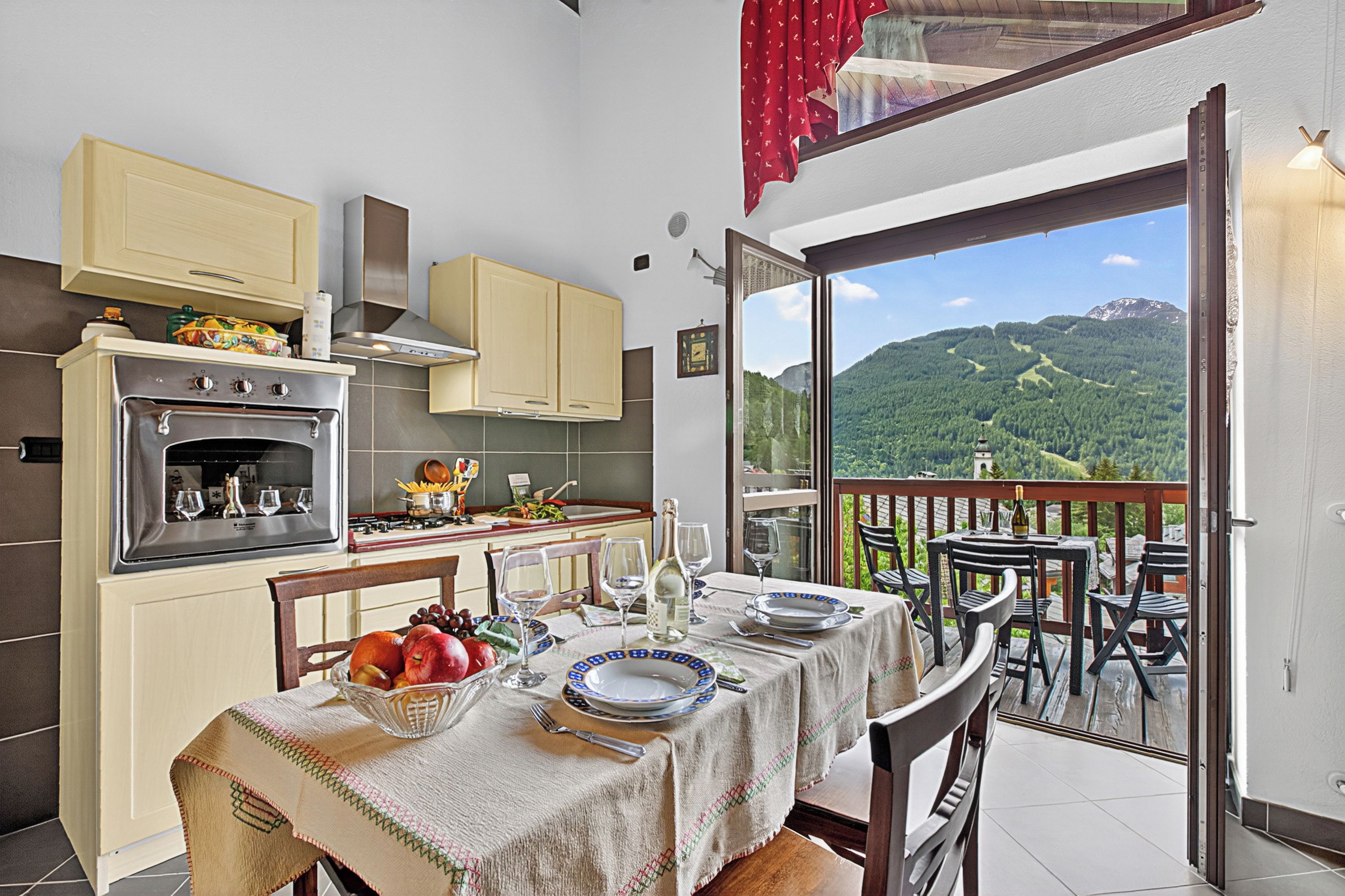 A kitchen with an oven, stove, and cabinets, a dining table set with plates and glasses, and a balcony with a mountain view.