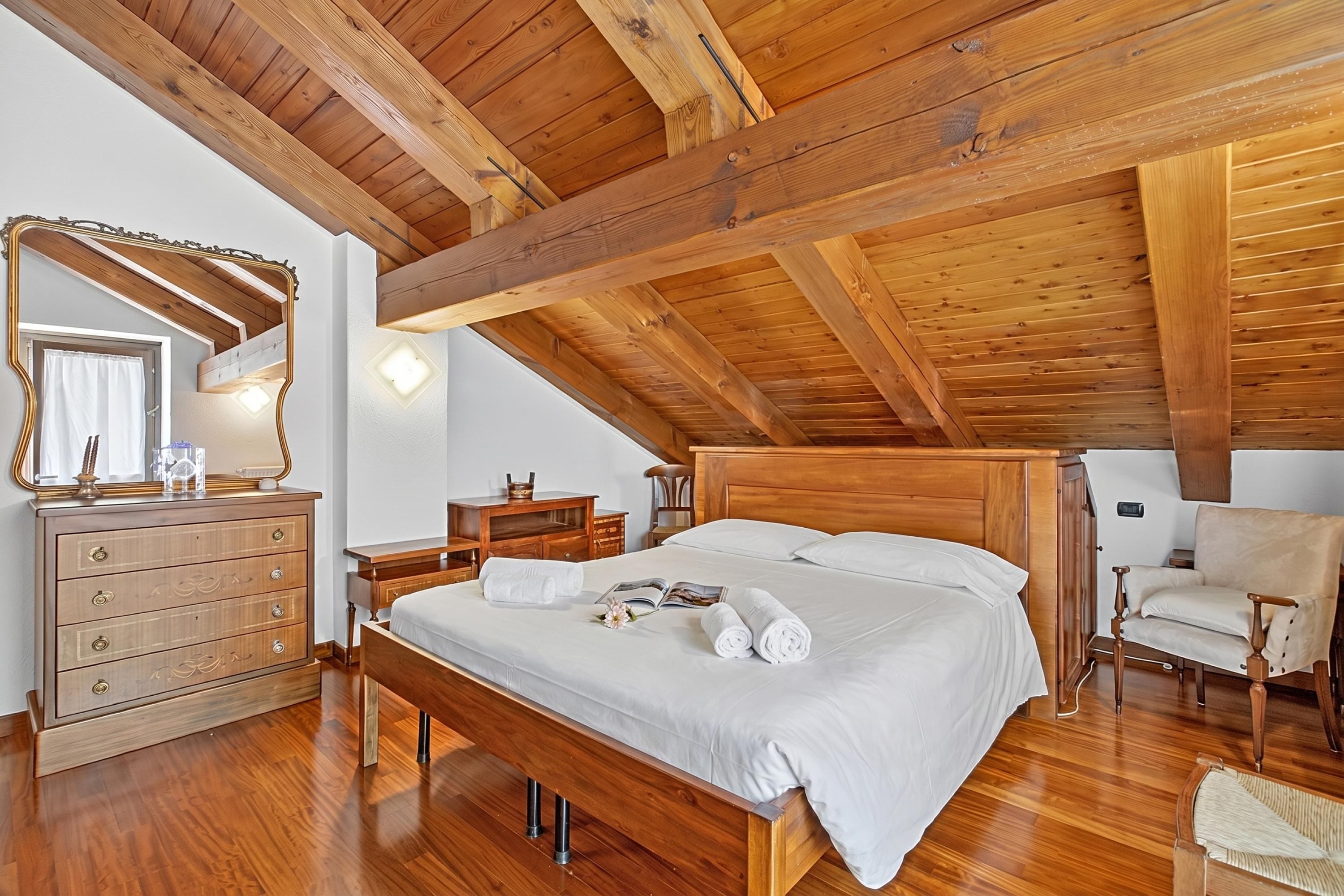 A bedroom with a wooden ceiling, a bed with white linens, a wooden dresser, a chair, and a mirror.