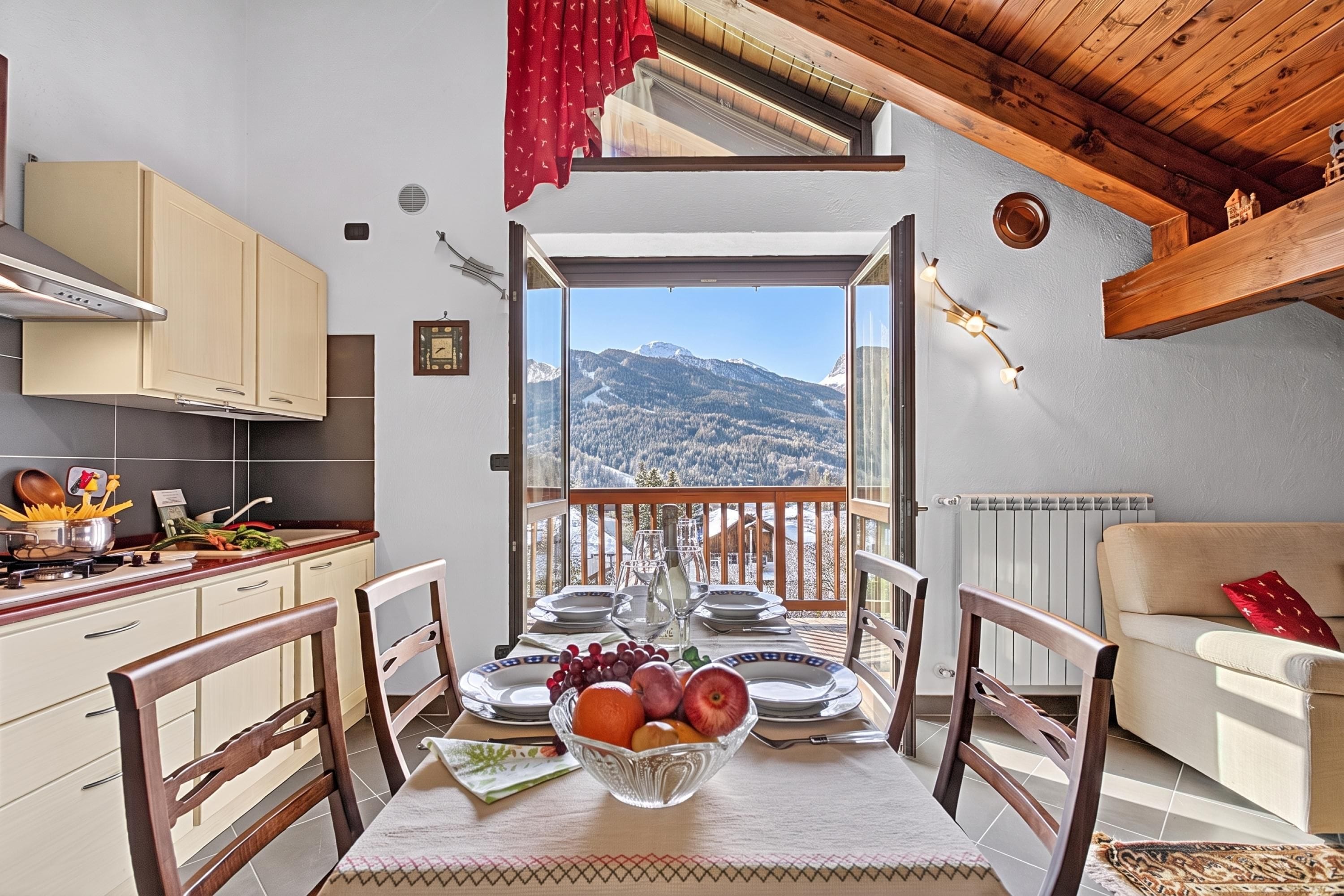 Un'area pranzo con un tavolo apparecchiato per due, vista sulle montagne e una cucina con elettrodomestici moderni.