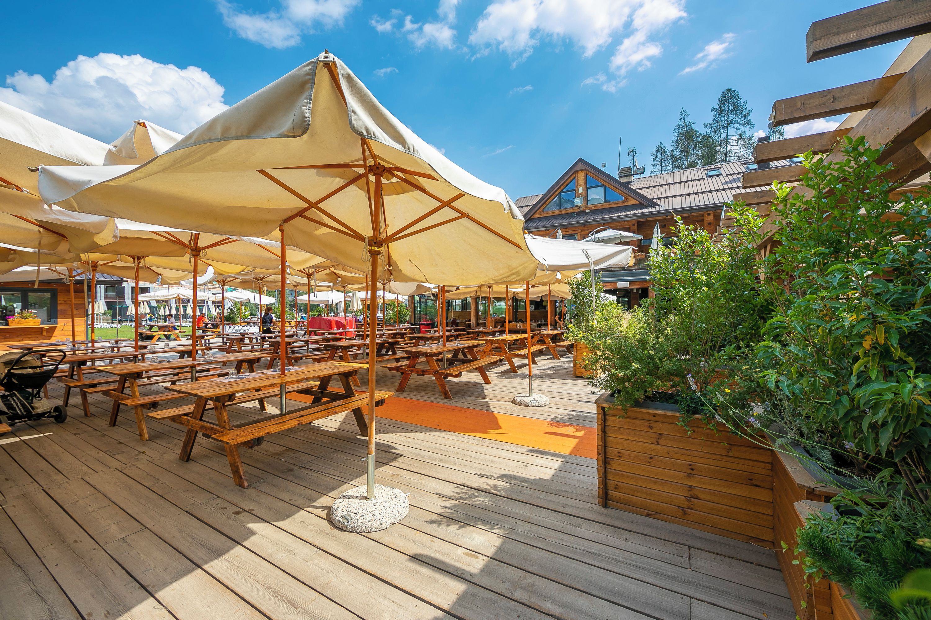 An outdoor wooden deck with picnic tables and umbrellas, surrounded by greenery and a building with a sloped roof.