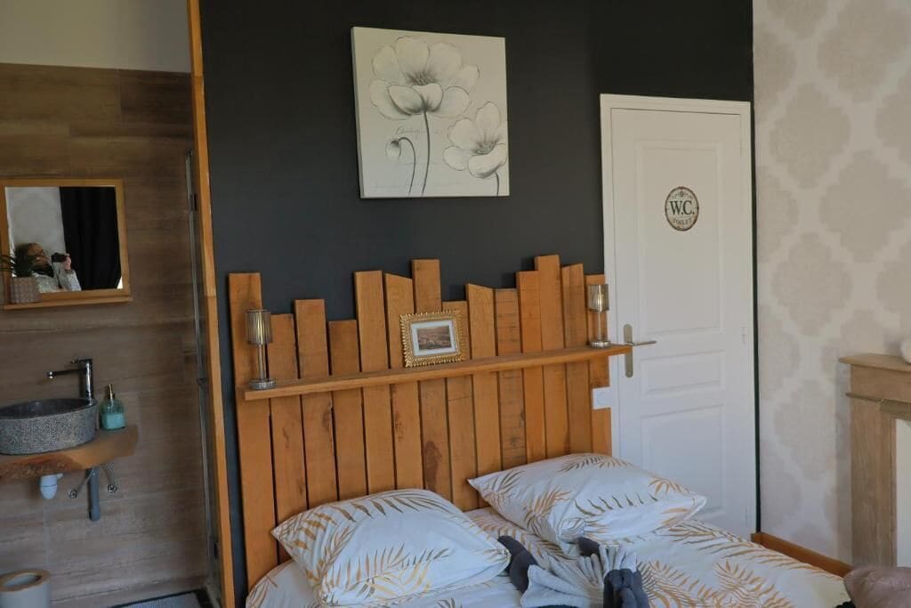 A bedroom with a wooden headboard, a bed with patterned bedding, a bedside table, a mirror, and a wall-mounted artwork.